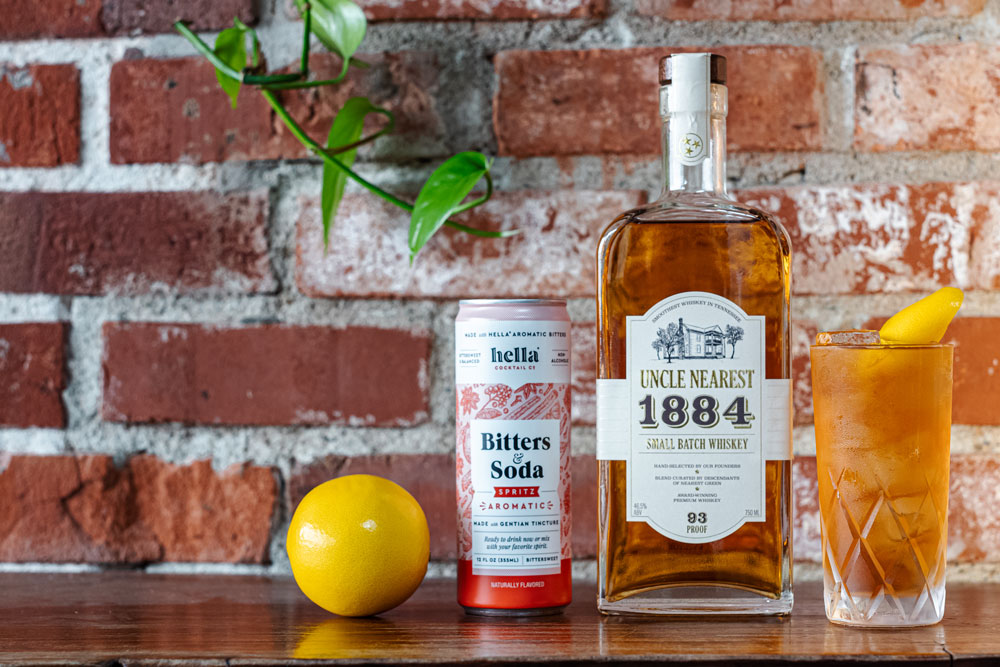 There is a brick background with an english ivy branch hanging down in front of it. Before it is a wooden table with a lemon, a can of hella bitters soda, a bottle of uncle nearest whiskey and the cocktail in a frosty glass. The cocktail is reddish orange and has a lemon peel garnish.