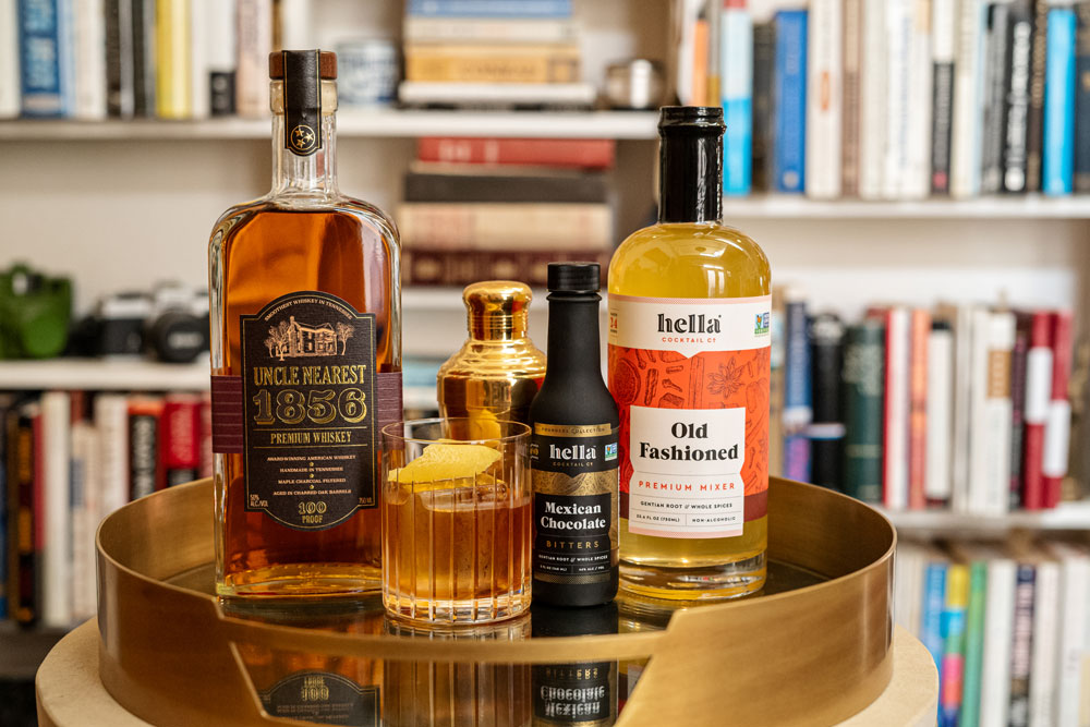 A blurry background of a bookshelf. In front is a gold circular serving tray with a bottle of uncle nearest whiskey, a gold shaker, a small black bottle of hella mexican chocolate bitters, a bottle of hella old fashion mixer, and a short glass of the cocktail with a lemon peel garnish.
